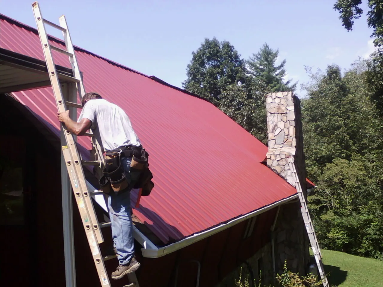 Roofer on a ladder beside a red metal roof and stone chimney — Soffit & Fascia Repair by Roofing Brothers in Santaquin